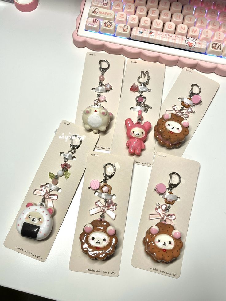 the image shows a collection of handmade keychains and a decorative keyboard. there are six keychains in total, each attached to a card that says "made with love". the keychains have a cute, kawaii aesthetic, featuring various animal characters (bear, bunny, cat) and food themes (cookie, rice ball). the keychains are adorned with beads, charms, and ribbons. a pink keyboard with decorative keys is visible in the background.