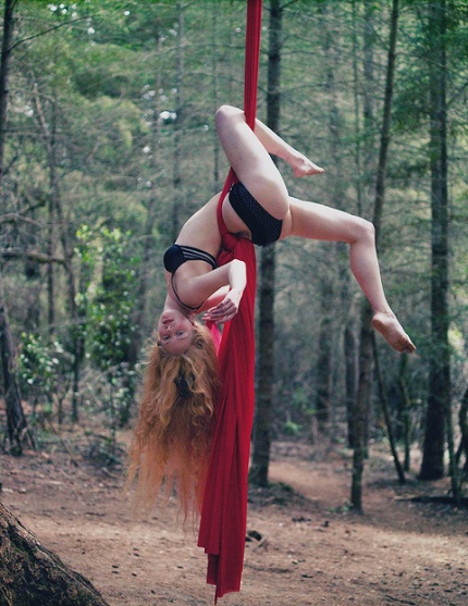 a woman is performing a beautiful aerial silks routine in the woods. she is suspended upside down, hanging from a red fabric. the forest around her is full of green trees. the woman's long, red hair cascades down her back. she is wearing a black bra and a black bikini bottom. the sunlight shines on her, giving her a golden glow. her arms are strong and flexible, holding the red fabric in place. she is in perfect balance and control, her body moving with the grace of a dancer.