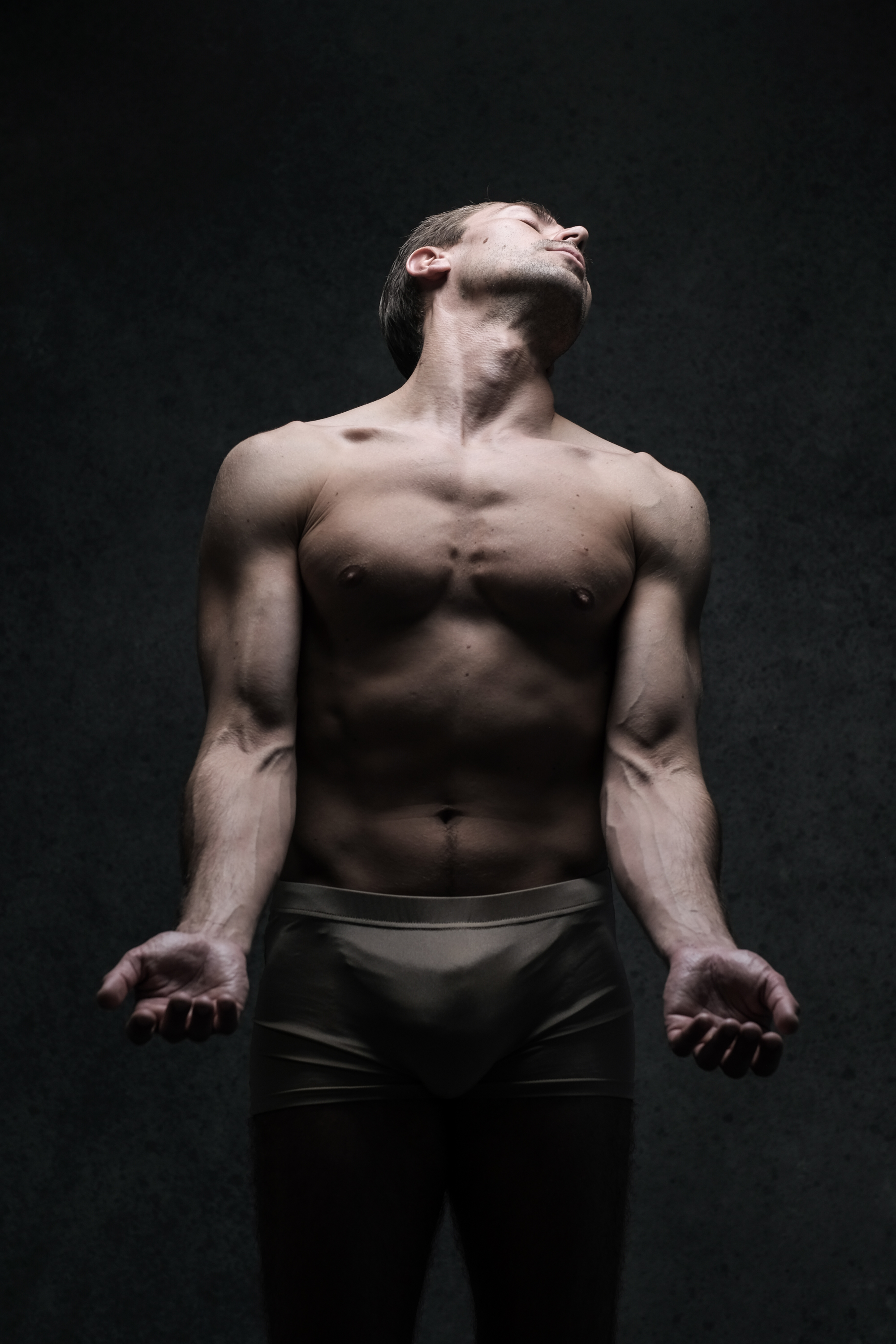 a shirtless man in tan underwear stands against a dark gray background, illuminated by a spotlight from above. his head is tilted back, eyes closed, arms outstretched with open palms facing upward in a pose that suggests surrender or vulnerability. his muscular physique is emphasized by the dramatic lighting.
