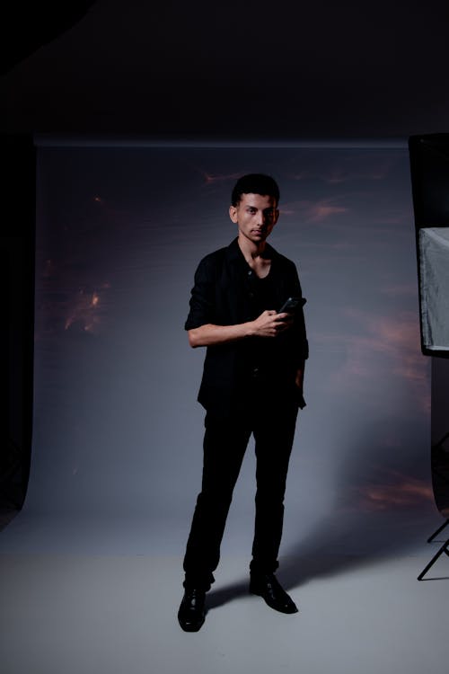 a young man in a black shirt and pants stands in a photo studio, holding a small device.  he is posing for a portrait shot. the studio background is a muted grey with subtle lighting effects. the overall mood is dark and serious.  a photography light stand is partially visible in the corner.