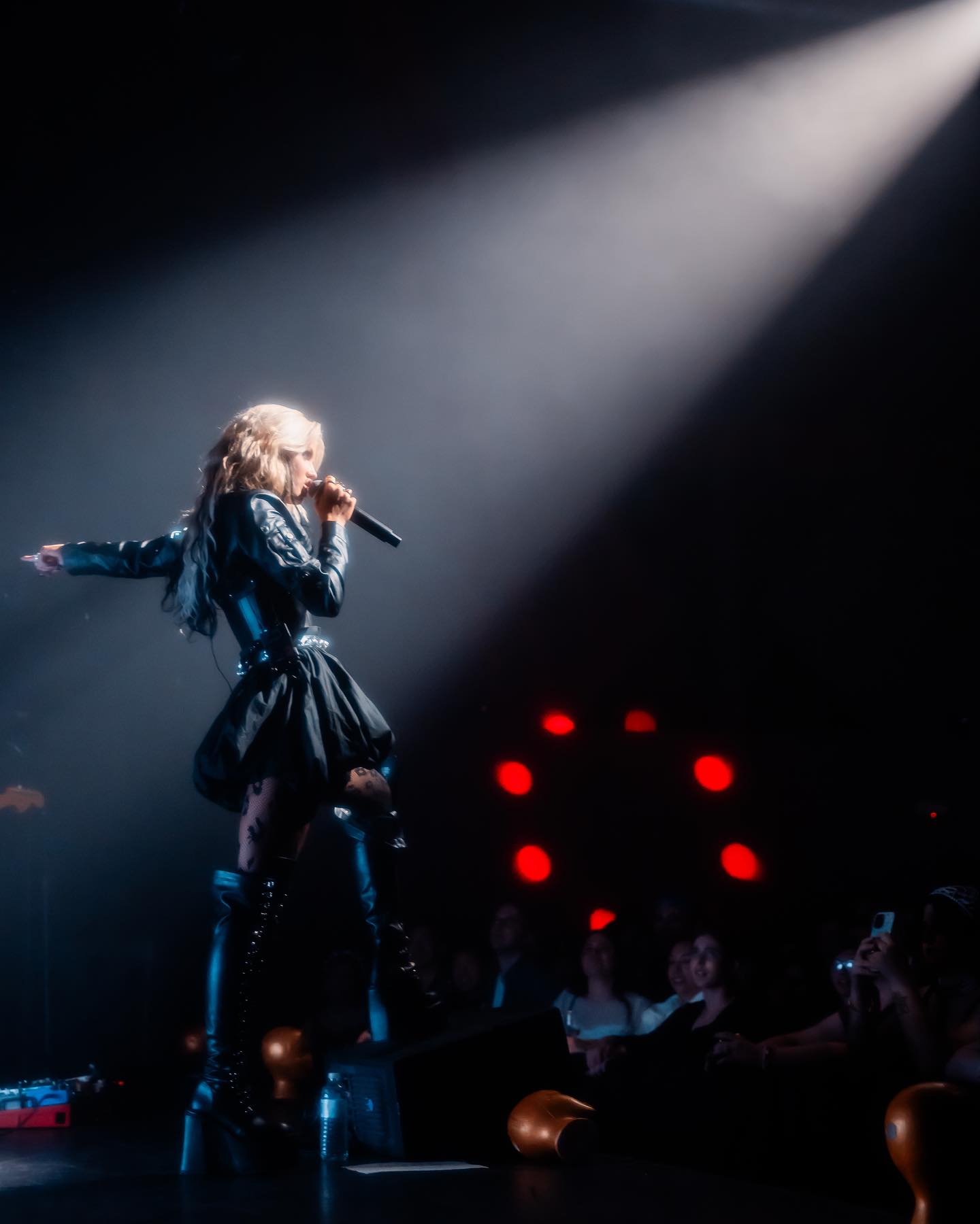 a singer performs on stage under a spotlight, holding a microphone and wearing black clothing including thighhigh boots, in front of an audience.