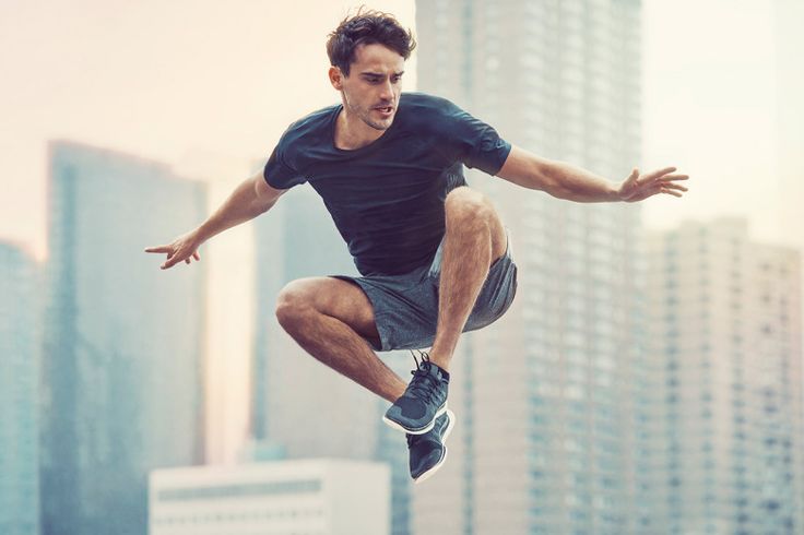a man wearing a black tshirt and dark shorts is in midair, jumping between buildings in an urban environment. the buildings are tall and sleek, reflecting the sun's rays. the man's expression is determined as he performs his athletic feat.  the image captures the energy and dynamism of urban life.