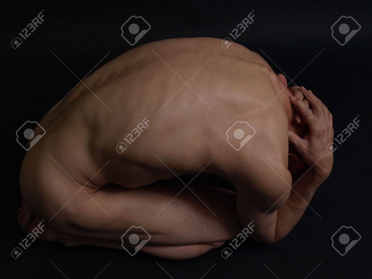 a nude male is curled up in a fetal position against a dark background. his head is down and his hands are holding his head or covering his ears. his skin is illuminated by the light in the room.