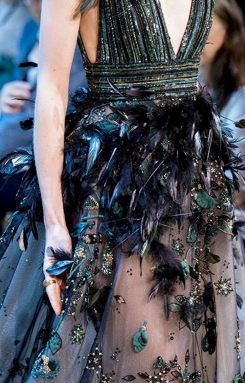 a woman in a blue and green dress with feathers and sequins. the dress is very elaborate and is designed for a special occasion. the woman's hand is in the foreground and she is wearing a ring on her finger. the image is of high quality and shows the details of the dress.