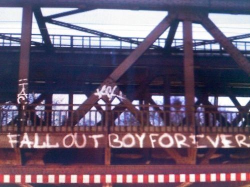 this image shows a closeup shot of a bridge in an urban environment. the bridge is made of steel and has a concrete base. there is graffiti on the bridge, which reads "fall out boy forever." the bridge is surrounded by greenery, suggesting that it is not abandoned. the image is taken from a low angle, giving the viewer a sense of perspective. the image is in focus and welllit.