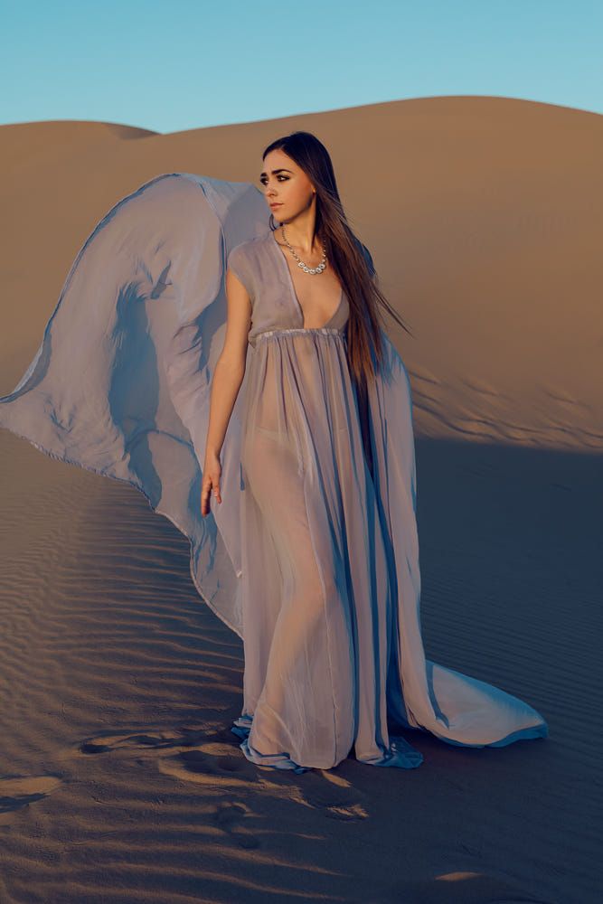 a woman in a flowing, sheer, lavender gown stands in the desert. the dress has a deep vneckline and a capelike detail that billows in the wind. she is wearing a silver necklace and has long brown hair. the sand dunes are a warm golden color, and the sky is a clear blue. the setting sun casts a warm glow over the scene.