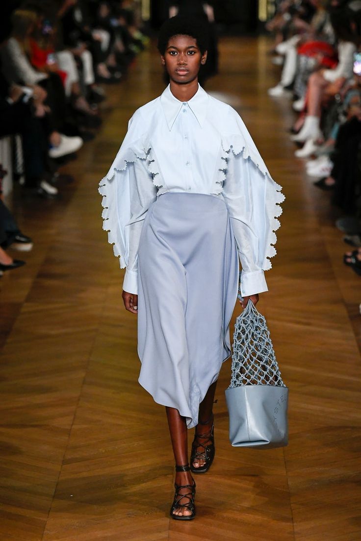 a female model walks down a wooden runway wearing a light blue, oversized shirt with ruffle detailing, paired with a matching midi skirt.  she's carrying a pale blue leather handbag with a net overlay.  the model is wearing black laceup sandals. the background shows other audience members. the overall setting suggests a fashion show.