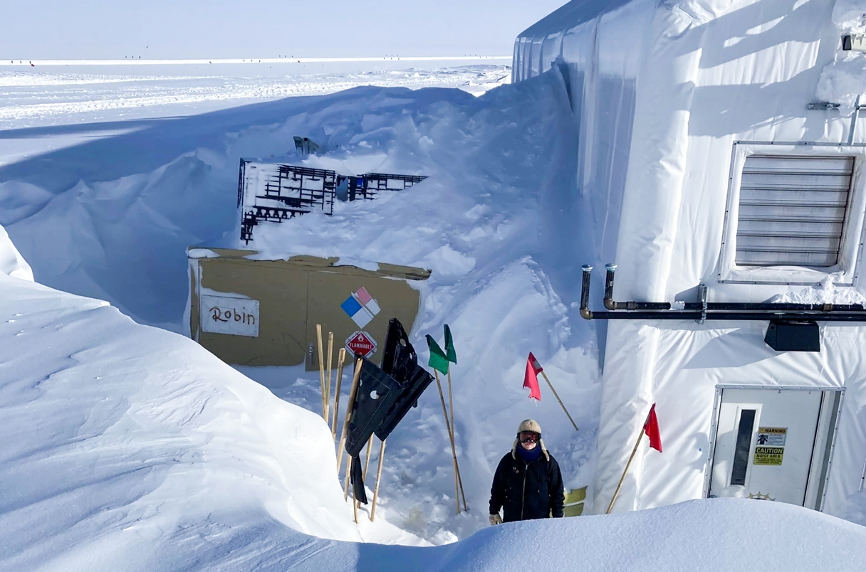 a person standing in front of a research station in antarctica. the station is made of a white fabric structure and is covered in snow. there are flags and equipment scattered around the station. the snow is deep and the landscape is white and frozen.