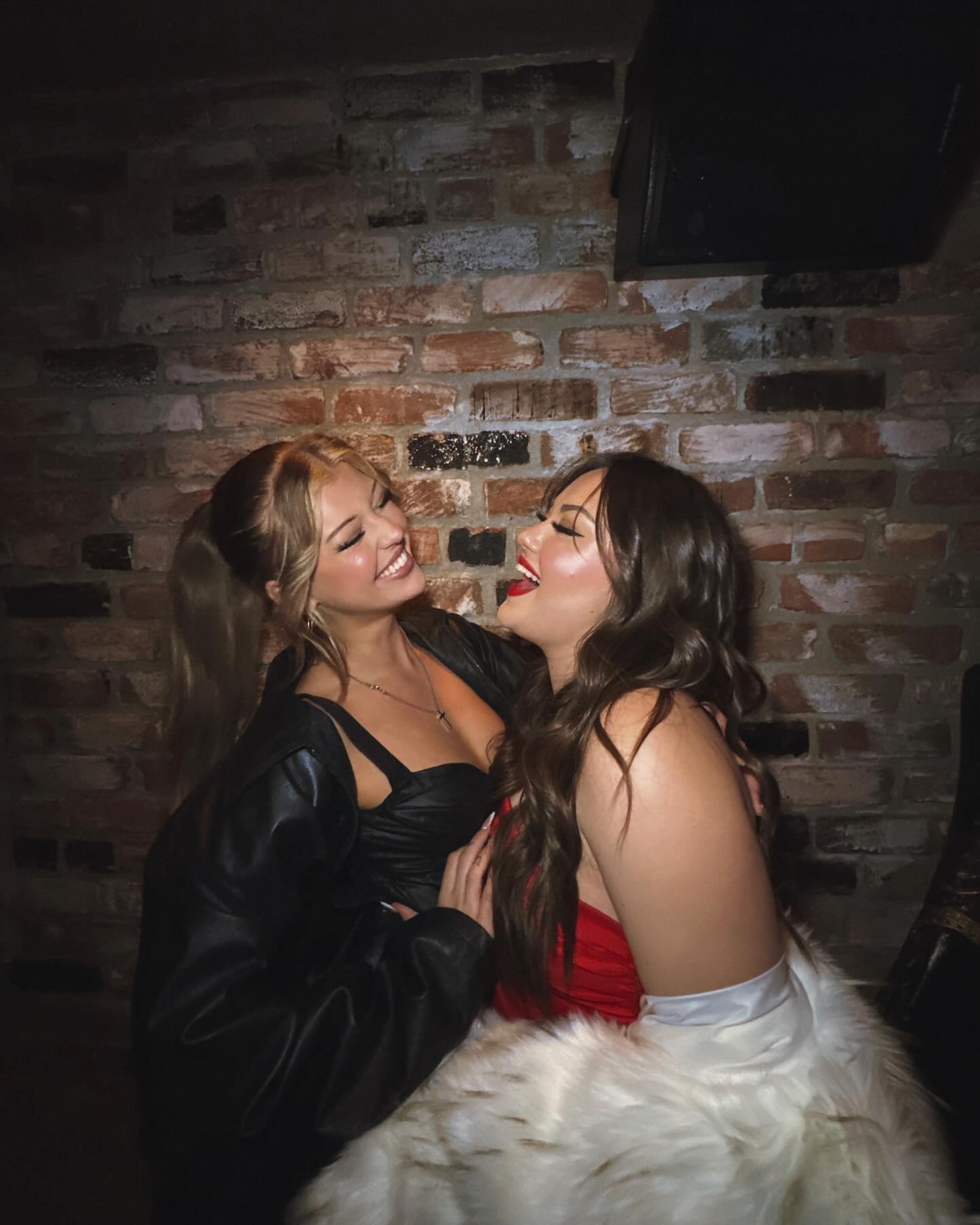 two women are posing indoors against a brick wall. the woman on the left has blonde hair in a high ponytail and is wearing a black leather jacket over a black strapless top. the woman on the right has brown hair and is wearing a red strapless dress with a white faux fur wrap. both women are smiling and appear to be enjoying each other's company.
