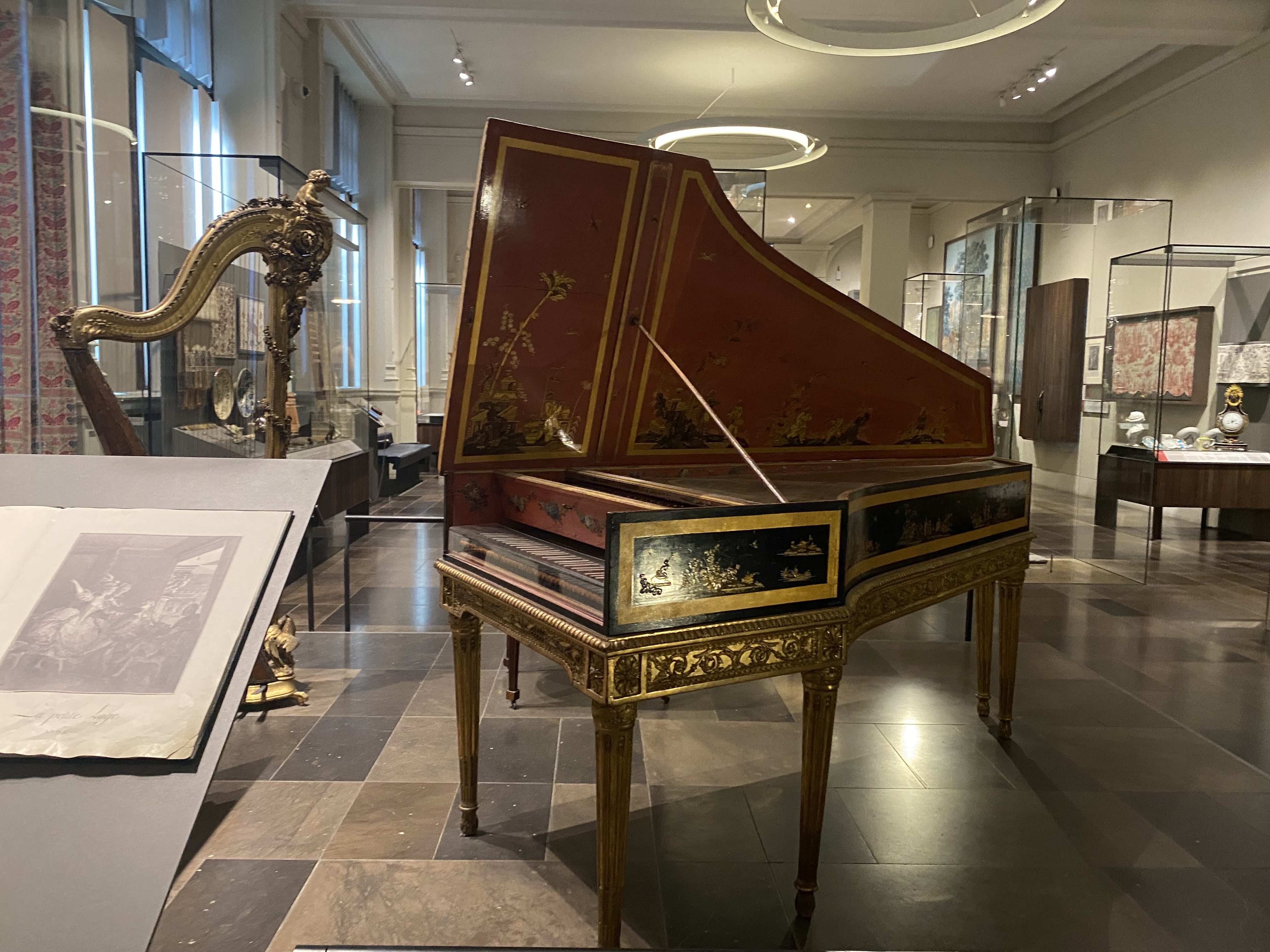the image shows a museum exhibit with several antique musical instruments and other artifacts. a red and gold harpsichord with its lid open is in the center of the frame. to the left is a gilded harp and a display with an open book. in the background, there are glass display cases containing various items. the floor is patterned with dark and light tiles. the lighting is provided by modern circular fixtures on the ceiling.