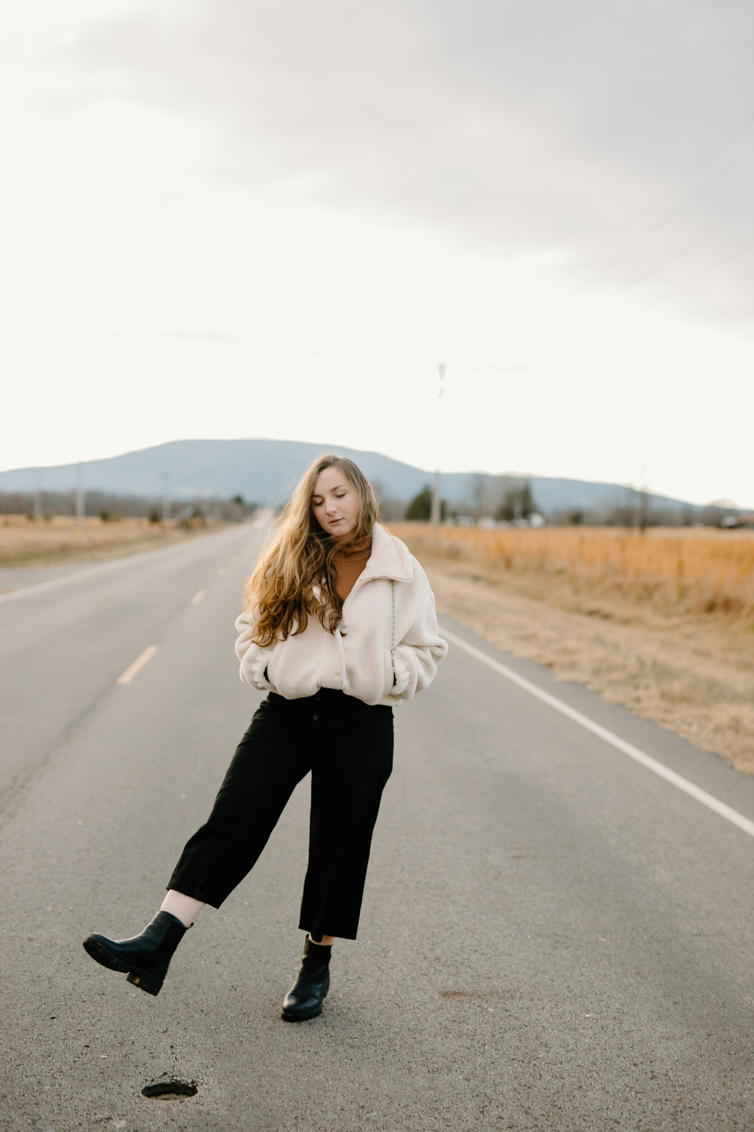 a young woman with long blonde hair stands in the middle of a rural road, wearing a creamcolored fluffy jacket, black wideleg pants, and black ankle boots. she is looking down, and her pose is relaxed and carefree. the setting is a wide open road with fields on either side, under a slightly cloudy sky. the overall mood is peaceful and serene.
