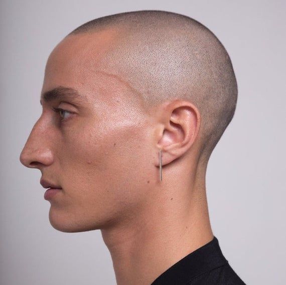 the image is a closeup, profile view of a young man with a shaved head. he has pale skin and is wearing a single, minimalist silver bar earring in his right ear.  his expression is neutral. the background is a plain, light gray, providing a clean and simple setting that focuses attention on the man's features. the overall style is minimalist and clean.