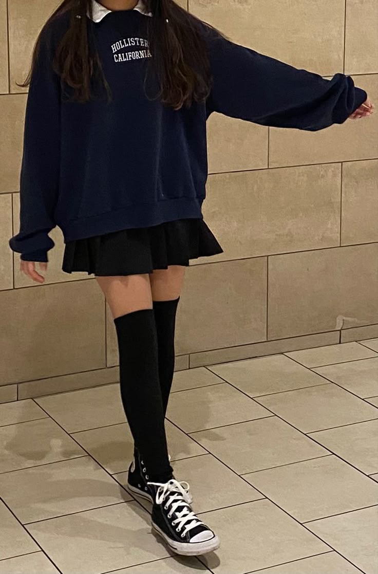 a person wearing a hollister california sweatshirt, a collared shirt, a black skirt, thighhigh socks, and black converse sneakers stands in front of a tiled wall and floor.