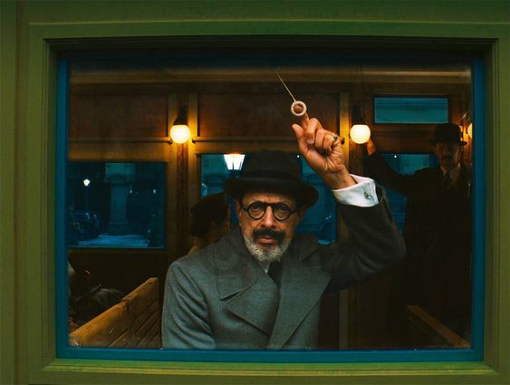 a man in a hat and glasses sits in a train car and looks out a window. he has a beard and mustache and looks serious. the train car is old and has a retro feel. there is another man in the background.