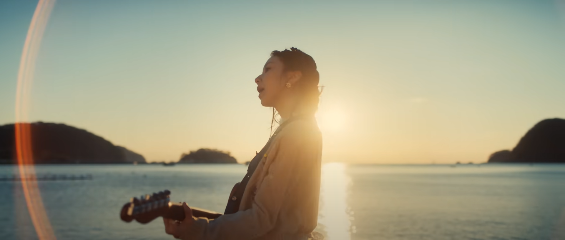 a woman stands on a beach at sunrise, silhouetted against the golden light, singing and playing guitar. the ocean and islands are visible in the background. the scene exudes a sense of peace and serenity.