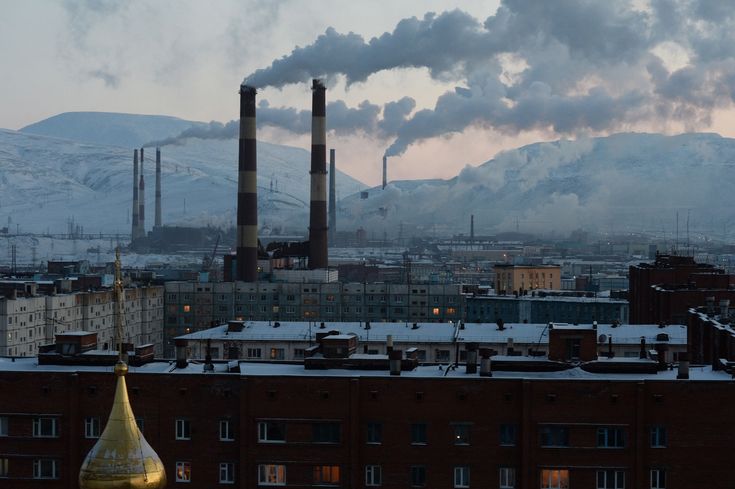 the image shows a city, possibly norilsk, russia, nestled in a snowy mountainous region.  dominating the scene are several large industrial smoke stacks belching thick plumes of dark smoke into the air, creating a heavy layer of pollution over the city.  the smoke obscures the background mountains somewhat.  the foreground consists of rows of residential buildings covered with snow, contrasting the industrial activity in the background. the overall mood is somber and emphasizes the environmental impact of the industrial complex.  a church dome is visible in the lower left corner, offering a small point of contrast against the industrial presence. the time of day seems to be either dawn or dusk, judging by the lighting.