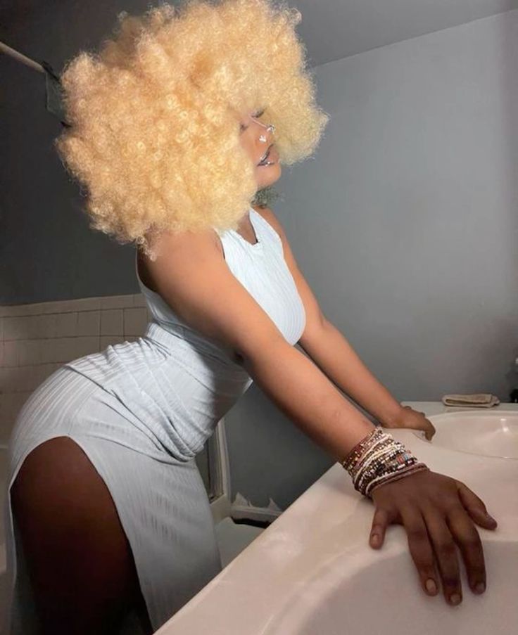 a young woman with blonde curly hair is wearing a white dress, standing in a bathroom, looking away from the camera. she is wearing many beaded bracelets and has her hand resting on the edge of a white sink.