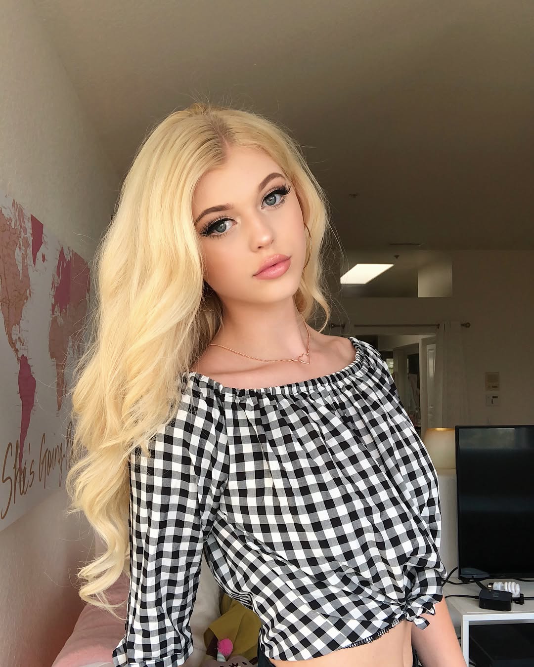 a portrait of a young blonde woman with fair skin wearing a black and white checkered offtheshoulder longsleeved shirt. she has long, wavy blonde hair, and is wearing makeup including eyeliner and lipstick. she is also wearing a delicate necklace and hoop earrings. behind her is a blurred background with a world map on the wall and a television on a table.