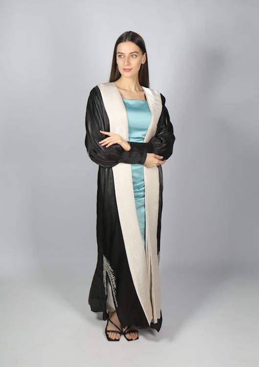 a woman in a black and white dress, standing in front of a gray background. the dress is longsleeved and features a light blue fabric underneath. she is wearing black highheeled sandals.