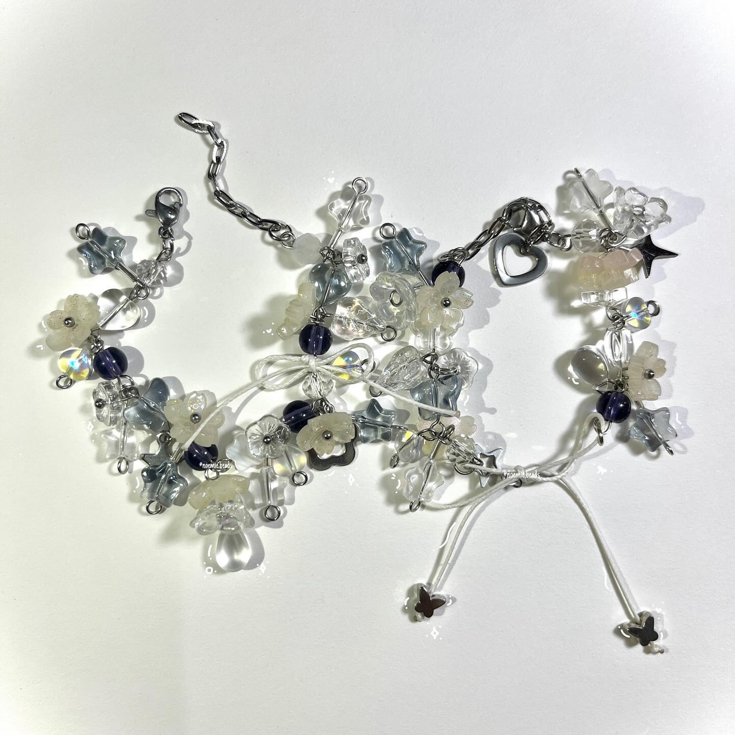 image shows two beaded bracelets on a white surface. the bracelets are made of various types of beads, including flower beads, star beads, mushroom beads, transparent beads, and purple beads. one bracelet has a metal chain with a lobster clasp, and a heart charm. the other bracelet has white string ends with butterfly charms. the bracelets are casting shadows on the white background.