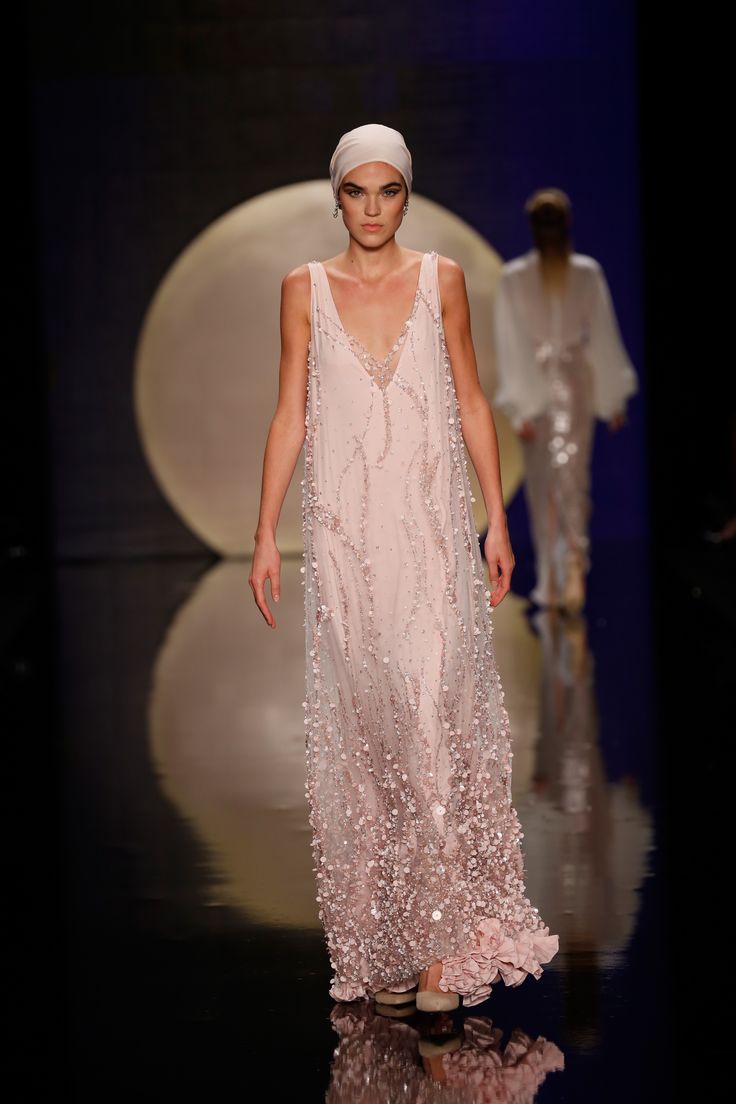 a model walks the runway at a fashion show, wearing a floorlength, pale pink evening gown with a vneck and sleeveless design. the gown is heavily embellished with beadwork and sequins, and features floral embellishments towards the hem. she also wears a matching headdress. the runway is highly polished, creating a reflective surface. in the background, there is a large, circular backdrop. another model is walking behind her.