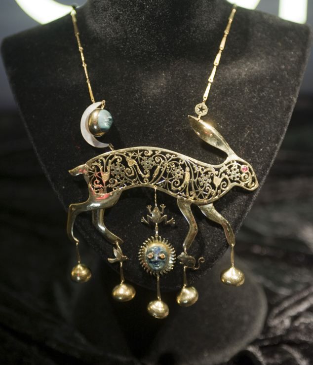 a detailed closeup shows an ornate, golden rabbit pendant necklace displayed on a black velvet mannequin. the rabbit, designed with intricate floral and geometric patterns cut out, features a red eye and various dangling charms, including golden balls, a sunlike face, and a small frog. the necklace also includes a crescent moon and a blue stone near the chain. the backdrop is a soft black velvet, enhancing the necklace's intricate details and luxurious feel.
