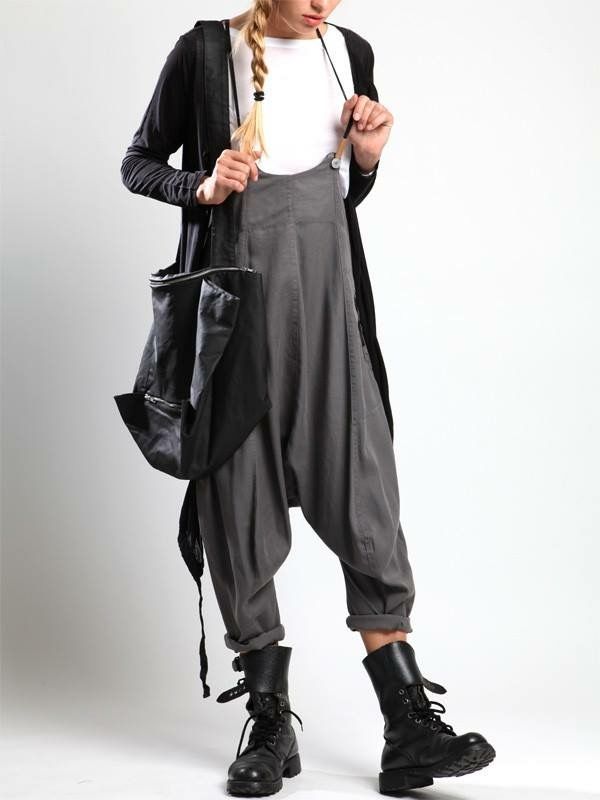 a woman is standing in front of a white background wearing a white tshirt, a black long sleeve cardigan, gray overalls, and black boots. she has a black bag on her shoulder. she has a blonde braided hair.