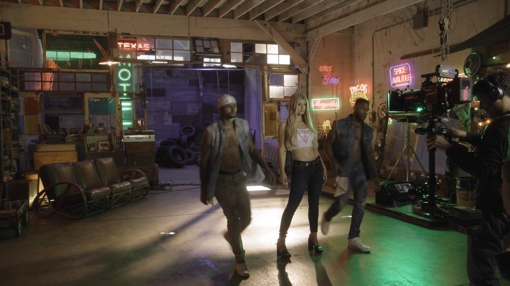 an indoor shot shows a film crew recording several models dressed in guess apparel in an industrialstyle setting. a man holding a camera is seen to the right of the shot, his face partially obscured by a headset. there is a blonde woman in the center wearing a cropped guess tank top and jeans. two men with open shirts and pants are to either side of her. behind them, there are many neon signs on the wall and to the left, a vintagelooking sofa.