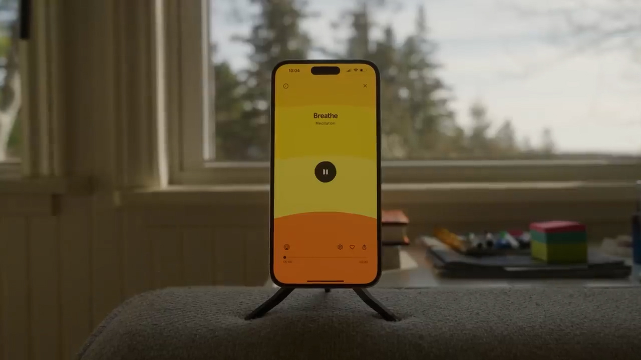 a smartphone is propped up on a small tripod on a grey surface, likely a couch or armchair. the phone displays a meditation app with a yellow and orange color scheme. the app is paused on a breathing exercise screen. through a nearby window, a blurry view of trees and a cloudy sky is visible. the overall lighting is soft and natural, suggesting daytime. the image evokes a sense of tranquility and peace.