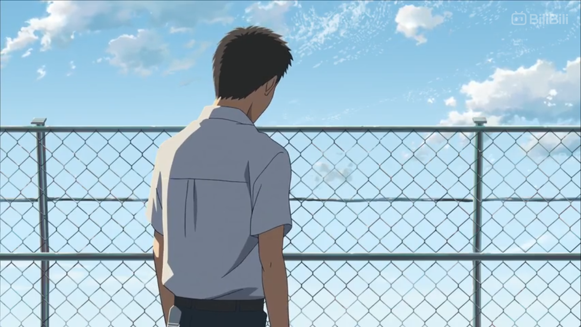 young man with short dark hair, wearing a white shortsleeved shirt and dark pants, stands facing away from the camera. he is looking out at a bright blue sky filled with fluffy white clouds. a chainlink fence stretches across the image, separating the man from the background. the scene gives off a sense of contemplation and solitude.