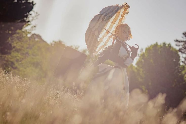 the image is a beautifully shot cosplay photograph of a woman dressed as violet evergarden.  she's wearing a detailed victorianera inspired dress and carrying a parasol.  the setting is a field of tall grass in the golden light of sunset or sunrise. the overall feel is dreamy, romantic, and evocative of violet evergarden's character and the show's aesthetic.  a suitcase is slightly visible in the background, further adding to the sense of journey and adventure. the photo is artistically styled with a soft focus and warm color tones.