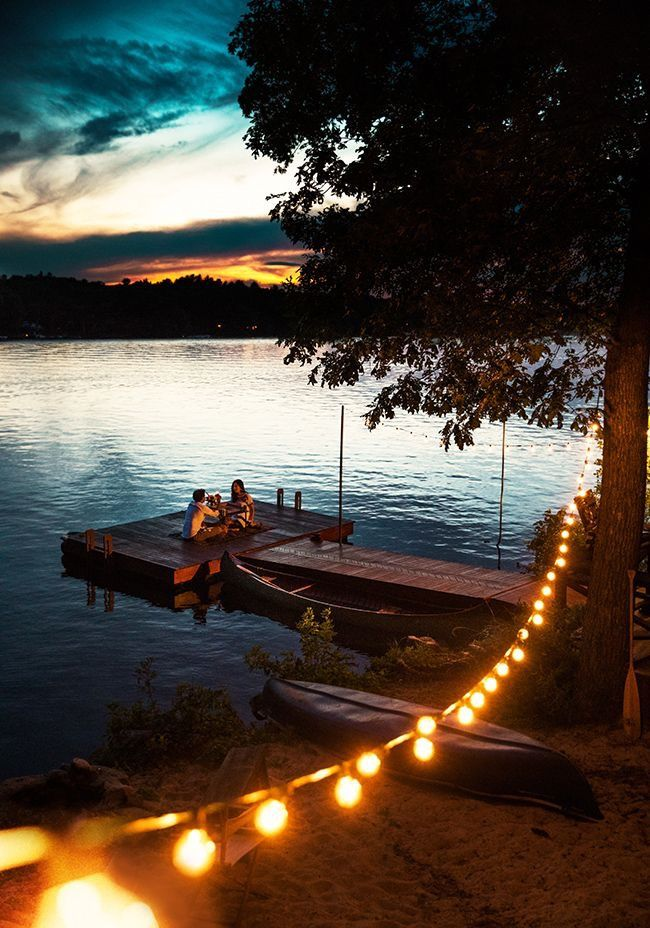a romantic scene unfolds at sunset on a lake. a couple sits on a dock illuminated by string lights, enjoying each other's company. canoes are moored nearby, and the tranquil water reflects the warm colors of the sky.  the scene is peaceful and idyllic, capturing a sense of intimacy and connection with nature.  the string lights add to the cozy and romantic atmosphere.