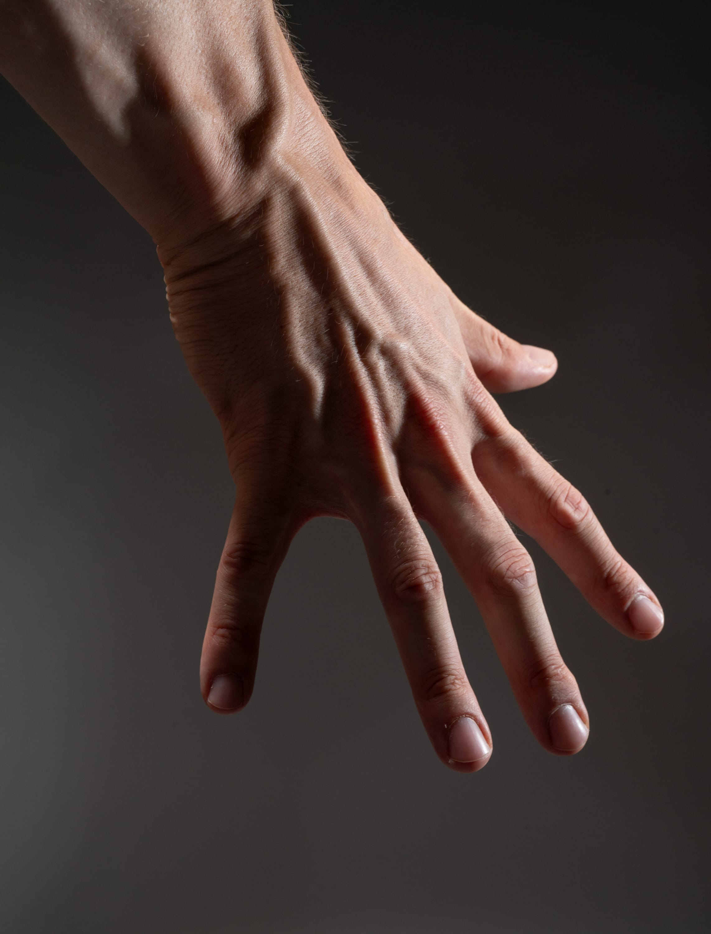 the image shows a man's hand extended against a dark background. the hand is muscular with prominent veins and tendons, illuminated by strong side lighting that creates dramatic shadows. the fingers are slightly spread apart in a relaxed gesture.