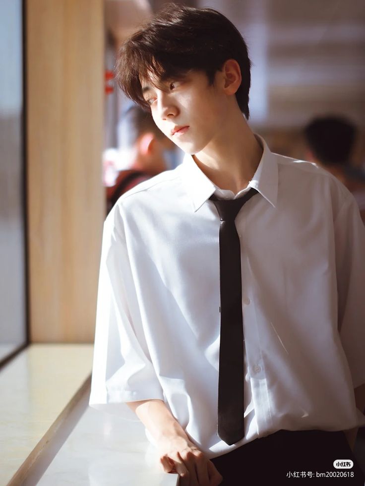 a young man, wearing a white shirt, black pants, and a black tie, stands by a window and looks to his right. he has short black hair and is leaning against the wall.  the lighting is bright, suggesting it's daytime, and the overall tone of the image is soft and warm.