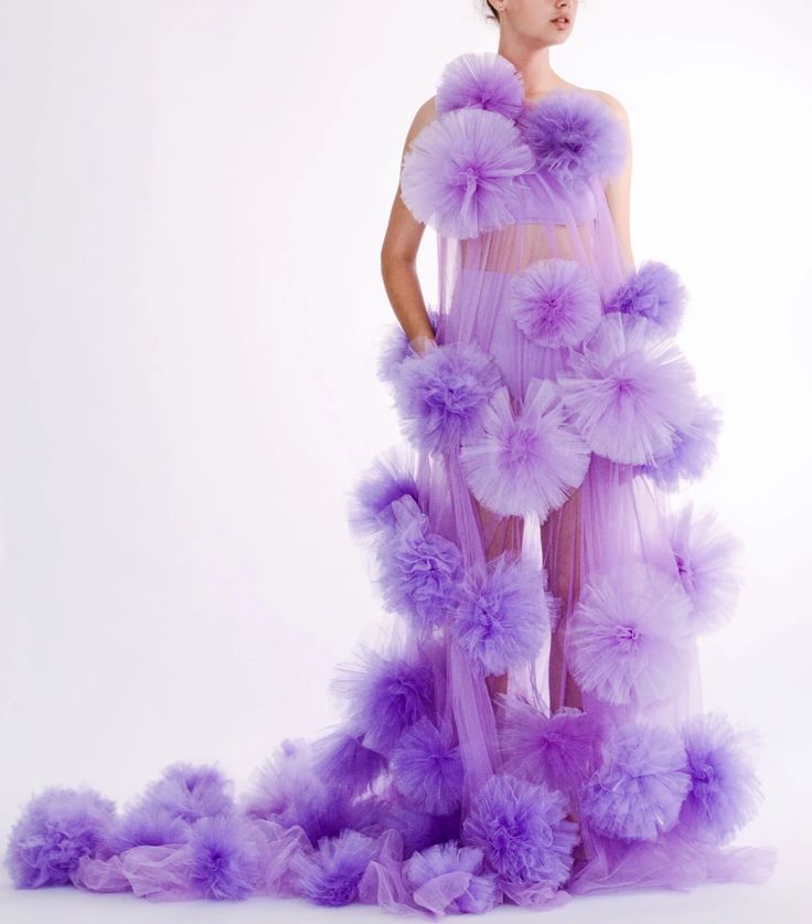 a woman in a purple tulle dress with fluffy pom poms on it, against a white background. the dress is sheer and elegant, making the woman look glamorous. the pom poms are arranged in a unique and creative way, adding a touch of whimsy to the outfit.  she is standing with her head slightly tilted to the side, and her hair is styled in a simple way. her makeup is minimal, and she looks confident and beautiful.