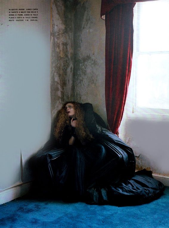a woman in a dark, long dress sits huddled in a corner. the room has a distressed wall, a red curtain, and a blue carpet. the overall aesthetic is vintage and somewhat melancholic.