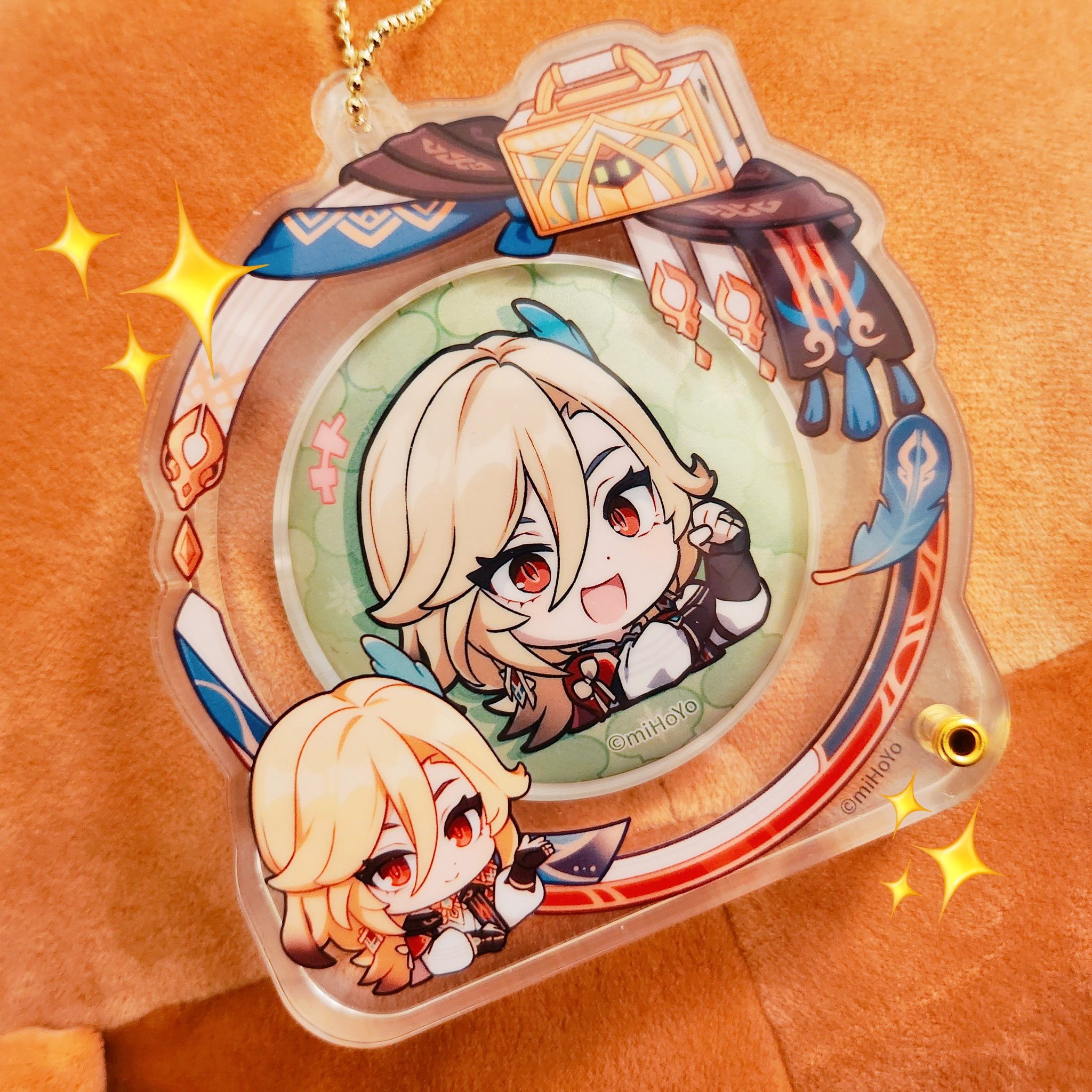a closeup photo of a genshin impact keychain featuring aether and lumine in chibi form. the keychain is made of clear acrylic and features a circular design with a colorful border. the background of the keychain is a light orange color, with glitter and sparkles scattered around the keychain.