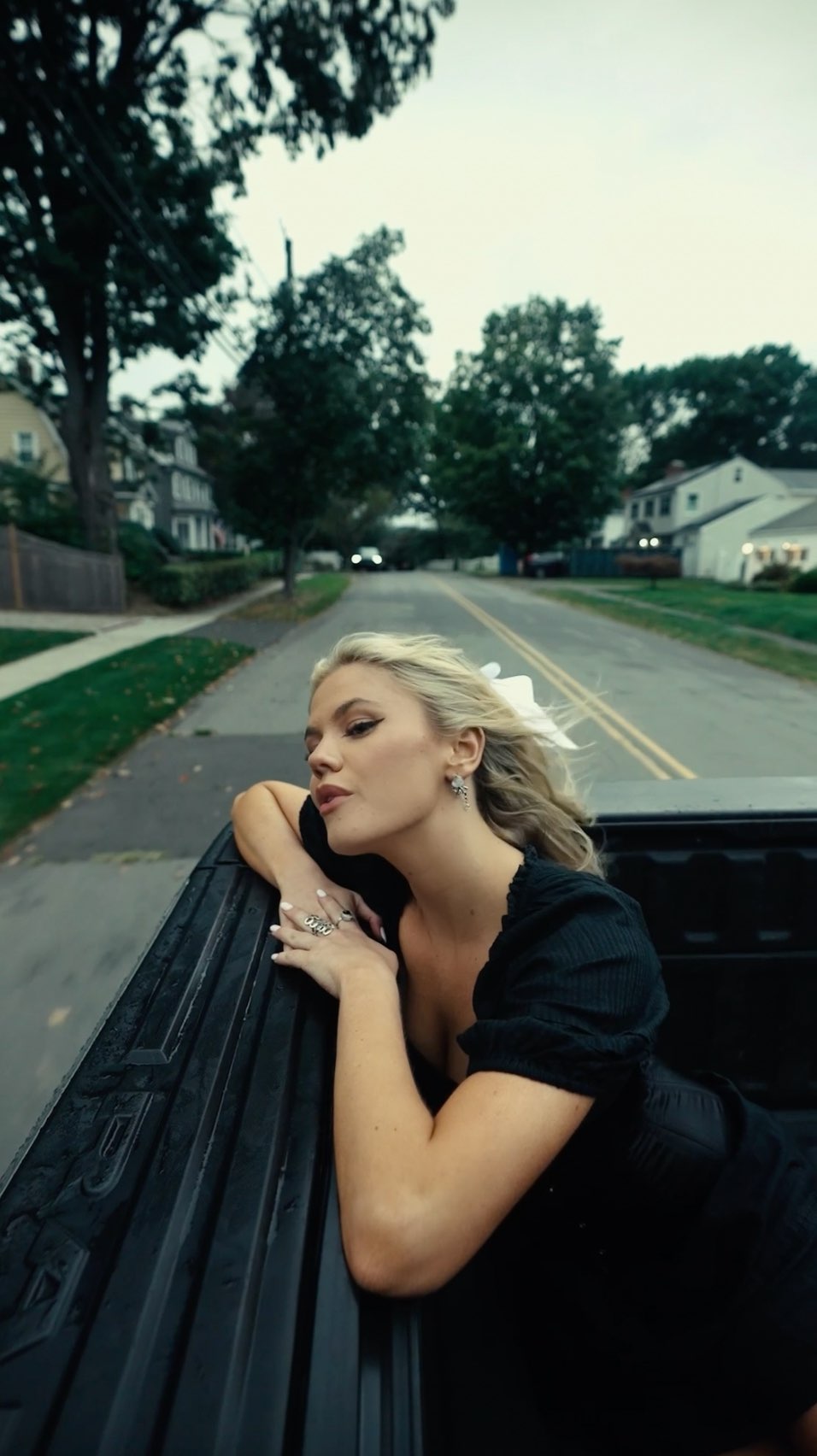 a blonde woman in a black dress is sitting in the back of a truck. the truck is driving down a road lined with houses and trees in a suburban area on an overcast day. the woman is wearing jewelry and a bow in her hair.