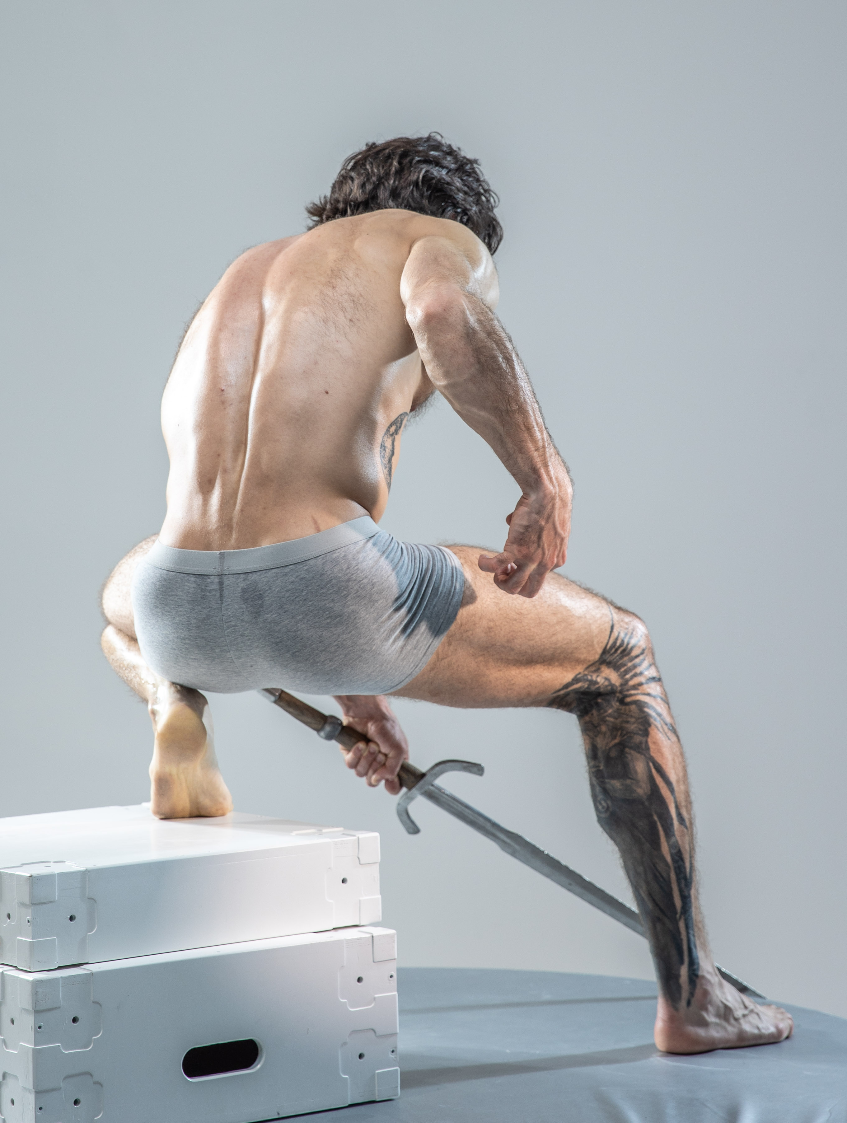 muscular man with tattoos on his leg and arm is captured in a dynamic pose against a simple gray background. he's crouching on a white platform, his back to the viewer, reaching for a sword that's lying in front of him. the studio lighting highlights the contours of his musculature.