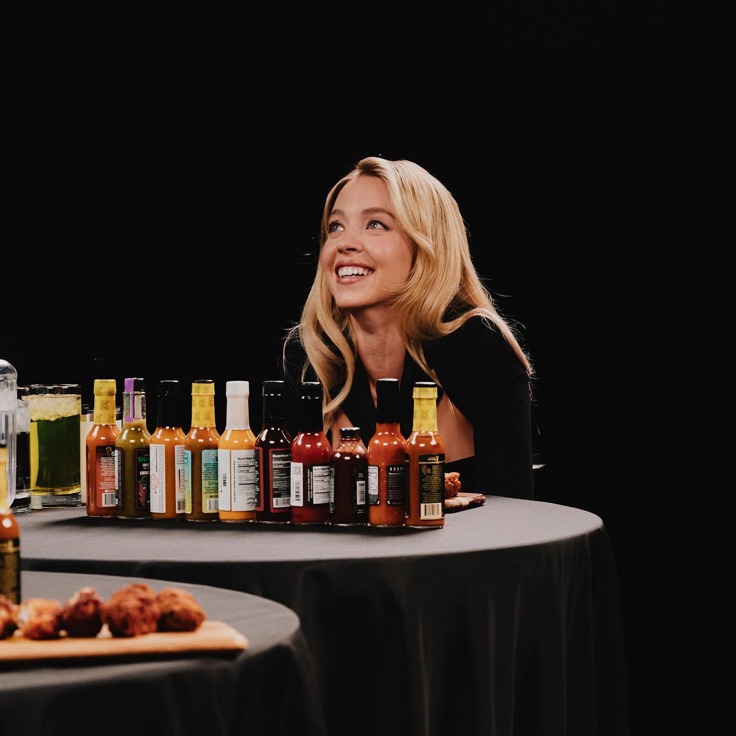 the image shows a young blonde woman with a bright smile, seated at a black table. in front of her is a row of various hot sauce bottles of different brands and colors.  there's also a small plate of chicken wings and a glass of a green beverage on the table. the background is dark and simple, suggesting a studio setting perhaps for a food show, interview, or product demonstration. the overall tone is bright and inviting, focused on the woman's happy expression and the array of hot sauces.