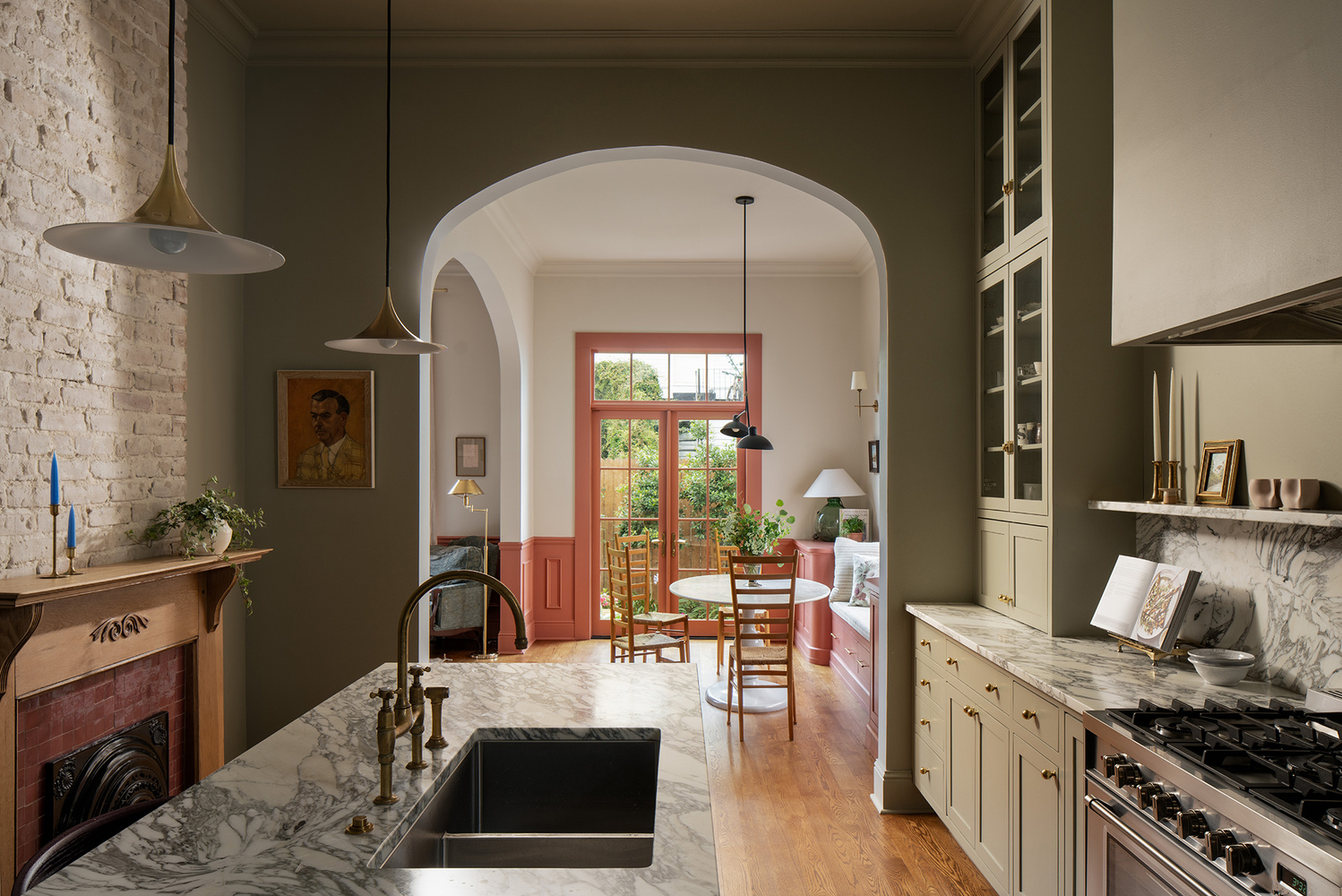 this image showcases a stylishly designed kitchen and dining area. the kitchen features marble countertops with a sink and faucet, complemented by custom cabinets and a stove. a brick wall with a fireplace adds a rustic touch, while artwork and unique lighting fixtures enhance the room's character. an archway leads to the dining area, which includes chairs, a table, and a window offering natural light. shelves display decor items, including a cookbook, candles, and a photograph. glass cabinets, wooden flooring, and a banquette complete the inviting and welldesigned space.