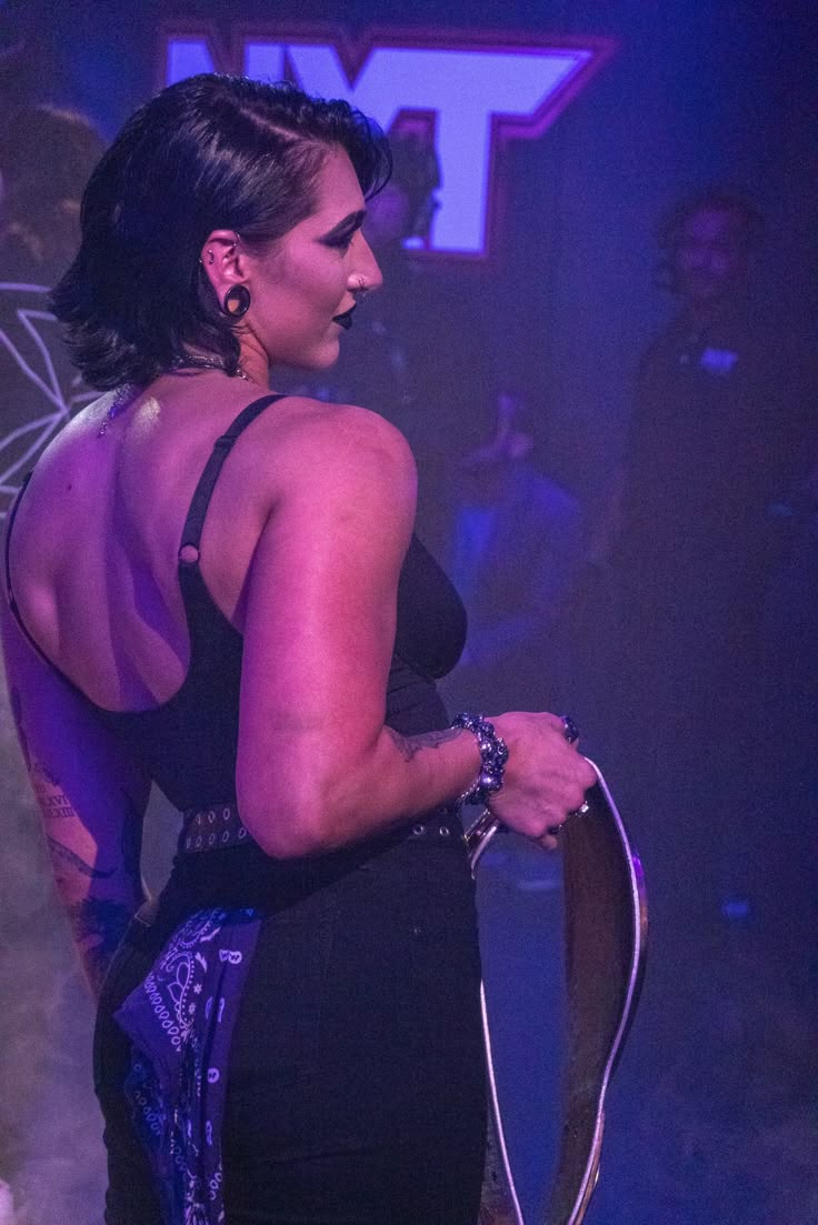a side view shot of rhea ripley, a professional wrestler, in a black outfit. she has dark hair and visible tattoos. she is holding a wrestling belt and wearing jewelry. a bandana is hanging from her outfit. the image is taken in the nxt environment.