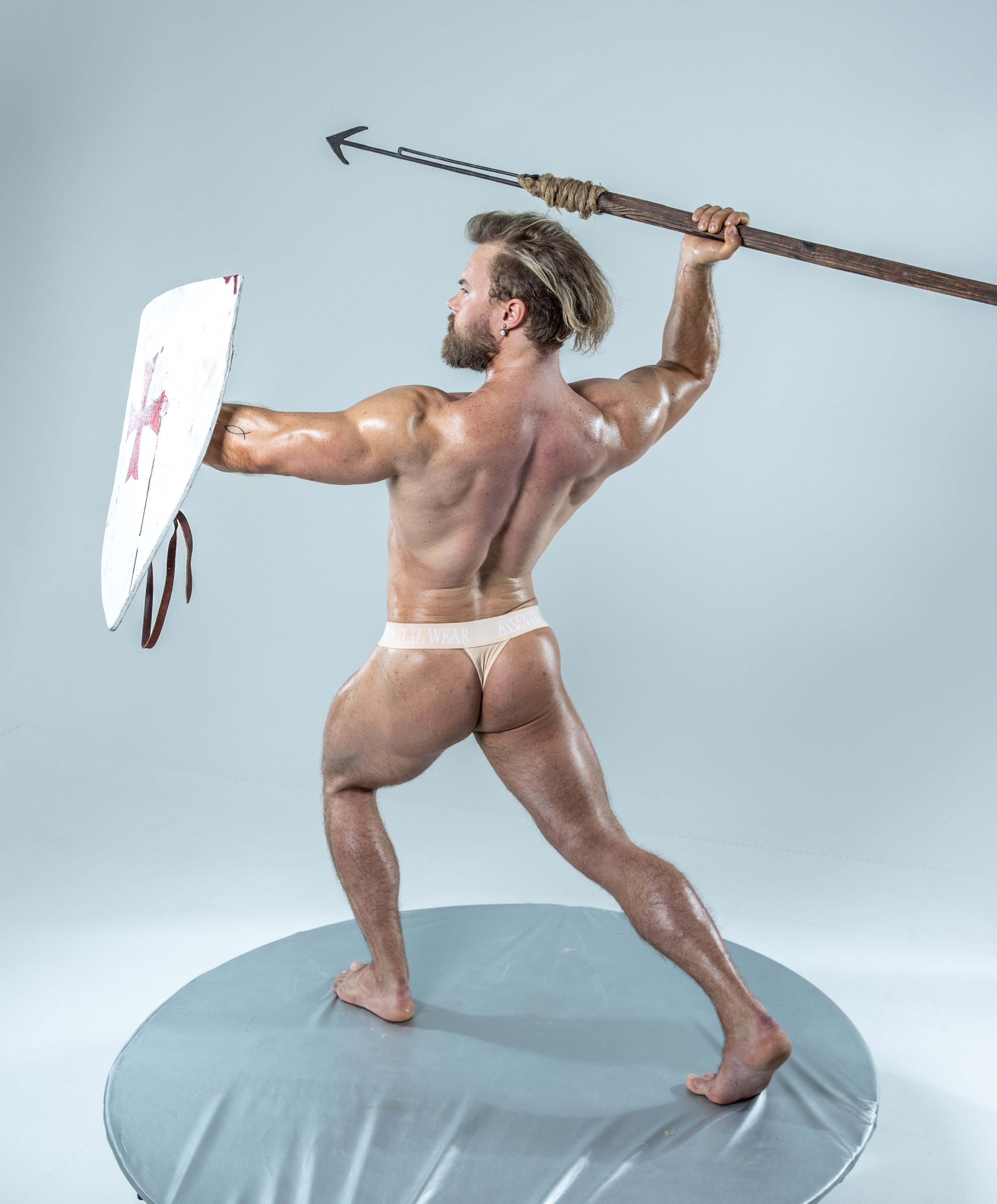 a muscular man stands on a grey circular platform against a plain grey background. he holds a spear aloft in his right hand, poised for action, and a shield in his left hand. he is nude except for a small beige loincloth. his physique is toned and muscular, suggesting strength and athleticism.  the scene is brightly lit, creating a dramatic and powerful image.