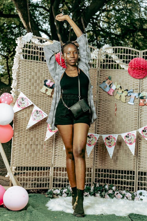 a young black woman stands smiling, arms outstretched against a wicker screen decorated for a birthday party. pink and white balloons, bunting, and flowers surround her.  she is wearing a grey cardigan, black shorts, a black top, a black handbag and ankle boots. the setting appears to be a park or outdoor space.  the overall mood is cheerful and celebratory.