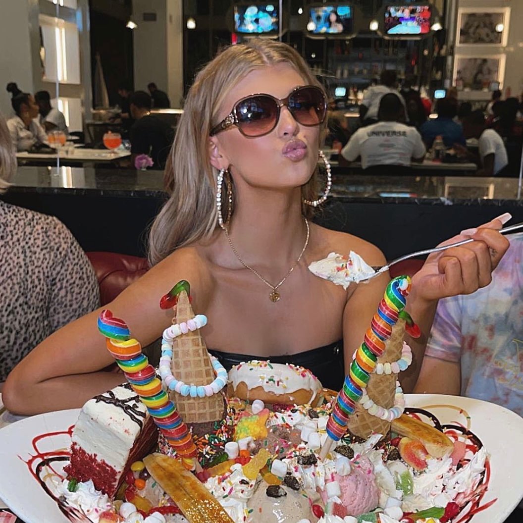 a woman with blonde hair, wearing sunglasses and hoop earrings, sits in front of a large plate piled high with ice cream, cake, lollipops, donuts, marshmallows, and sprinkles. she is holding a spoon with ice cream on it and making a kissing face. there are other people dining in the background.