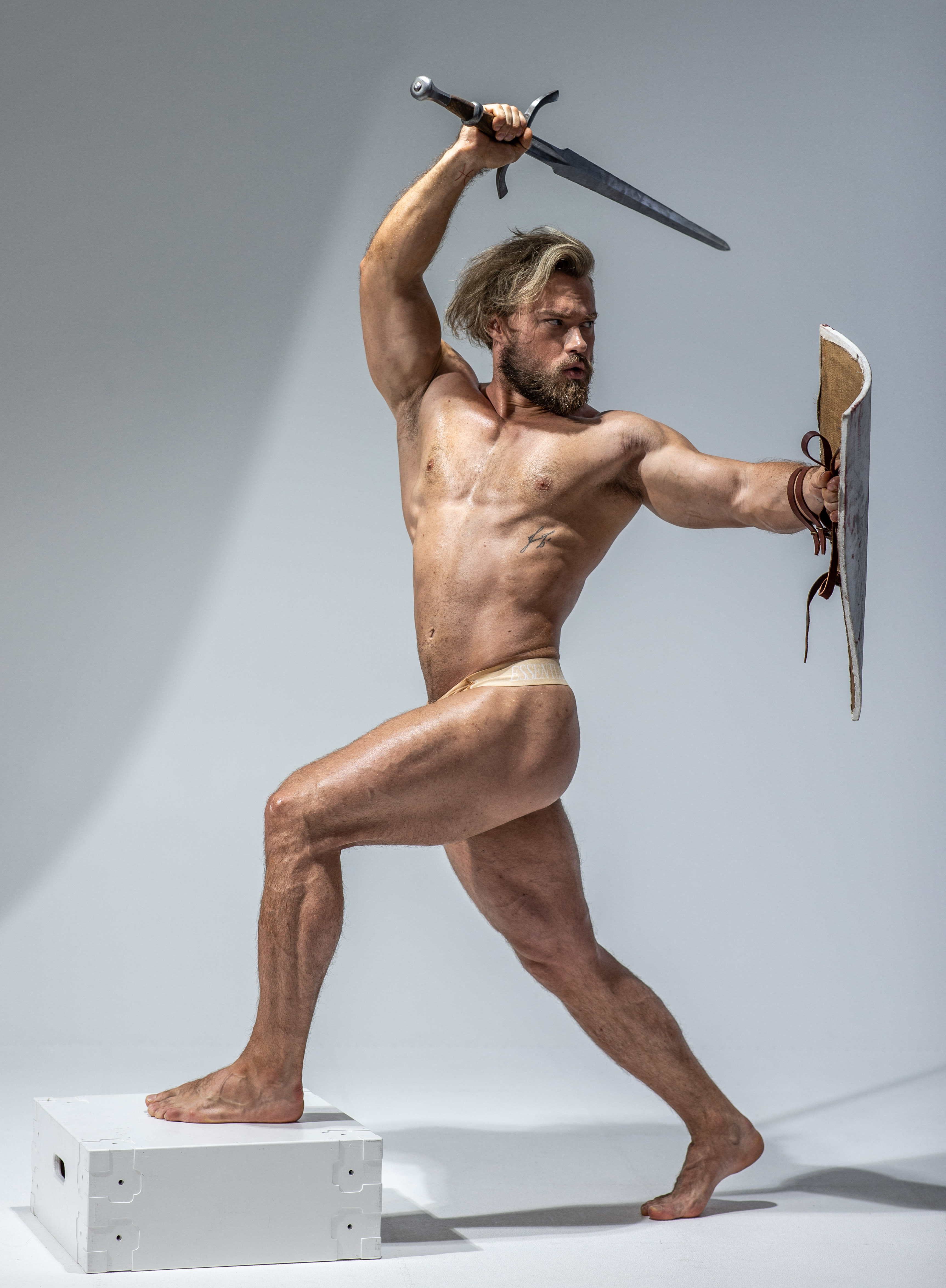 a muscular, nude man with long blond hair and a beard stands on a white platform against a white background. he adopts a dynamic pose, holding a sword raised above his head in his right hand while his left hand grips a large, leatherbound shield at his side. the image evokes themes of strength, power, and masculinity. the studio lighting highlights the definition of his musculature and creates a dramatic effect.