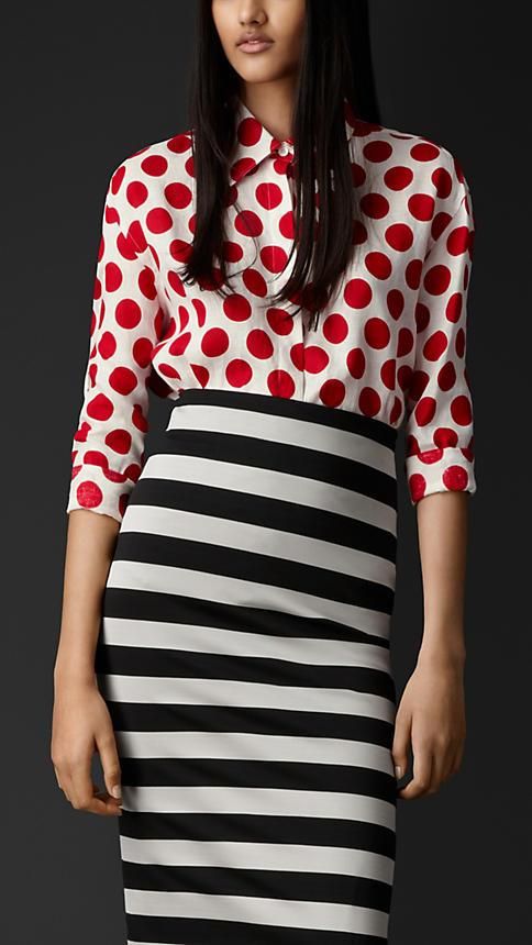 a woman is modeling a red polka dot shirt paired with a black and white striped pencil skirt. the shirt is longsleeved with a classic collar and buttondown closure. the skirt is a midi length, cut to hug the figure.  the model's dark hair is styled simply. the background is a dark gray, providing a clean and sophisticated aesthetic.  the overall style is elegant and contemporary.