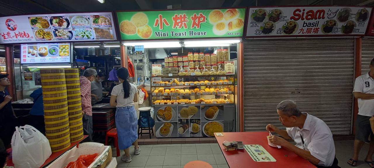 a bustling hawker center in singapore, showcasing the vibrant street food scene.  the image features a brightly lit food stall, "mini toast house," specializing in asian pastries and desserts. the stall is adorned with traditional chinese characters and boasts a variety of sweet treats displayed in glass cases. the image captures the lively atmosphere of the food court, with patrons enjoying their meals and vendors preparing fresh delicacies. the image also showcases other food stalls like "siam basil" and "生意小吃," offering a diverse culinary experience. the vibrant colors, enticing aromas, and bustling activity capture the essence of singapore's renowned hawker culture.