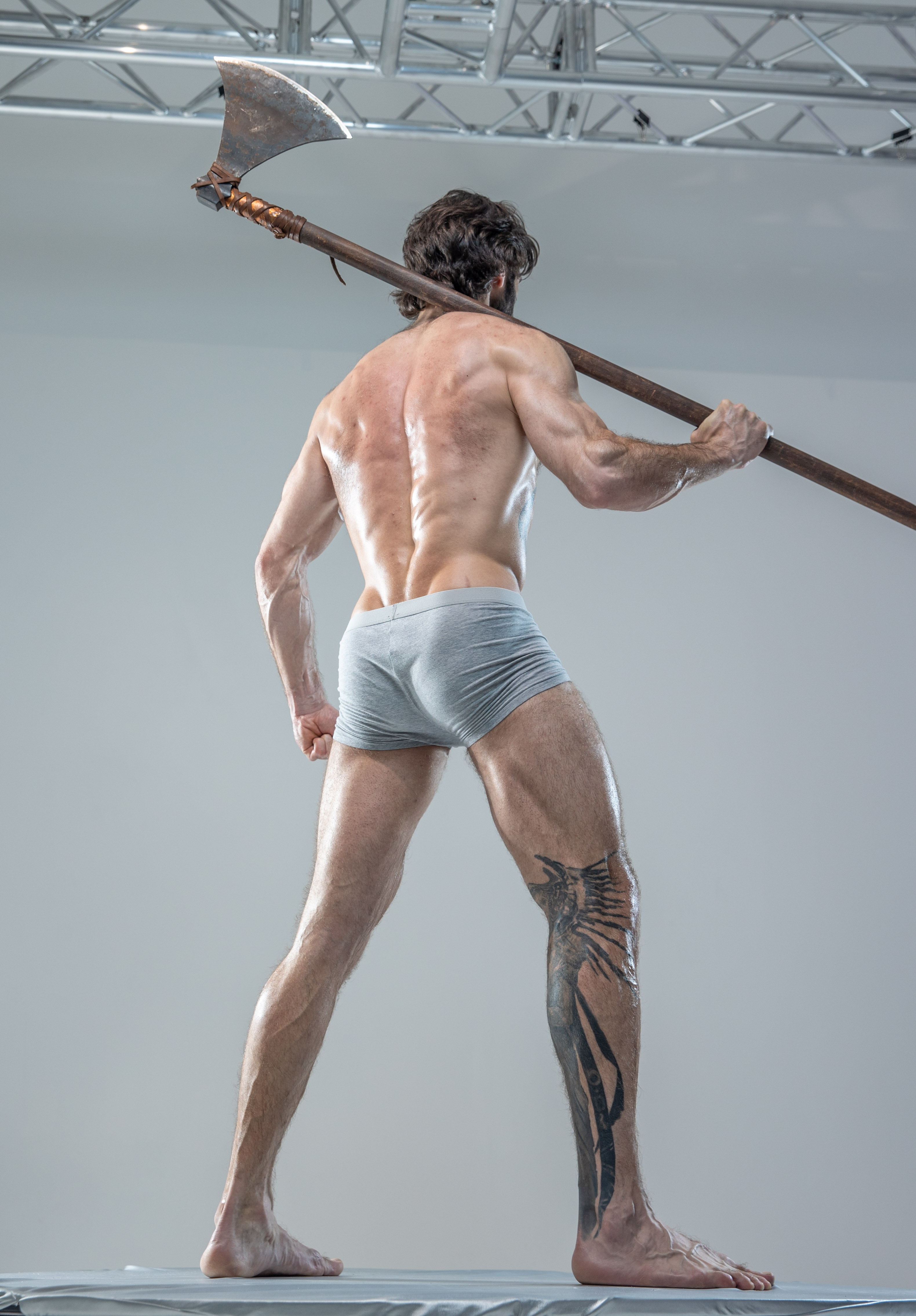 a muscular man with a tribal tattoo on his left leg stands with his back to the camera. he holds a large axe over his shoulder, its handle resting against his upper back. he is wearing only underwear, showcasing his physique. the setting appears to be a studio, with a plain grey background and soft, even lighting. the low camera angle emphasizes his height and powerful stance.