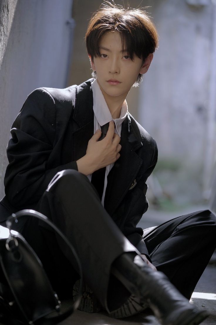 a young man with short, dark hair and a stylish, androgynous look is captured in an urban setting. he sits on a step, wearing a black suit jacket, white shirt, and black trousers. his gaze is directed slightly away from the camera. a black bag rests beside him. the image is illuminated by natural light, casting soft shadows and creating a natural, candid feel.